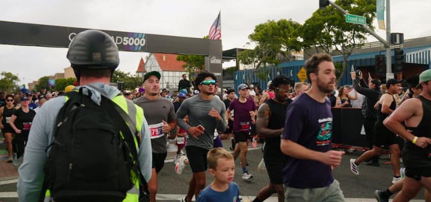 Runners participating in a crowded 5K road race, passing the starting line on a city street under an overcast sky, with spectators cheering along the course.