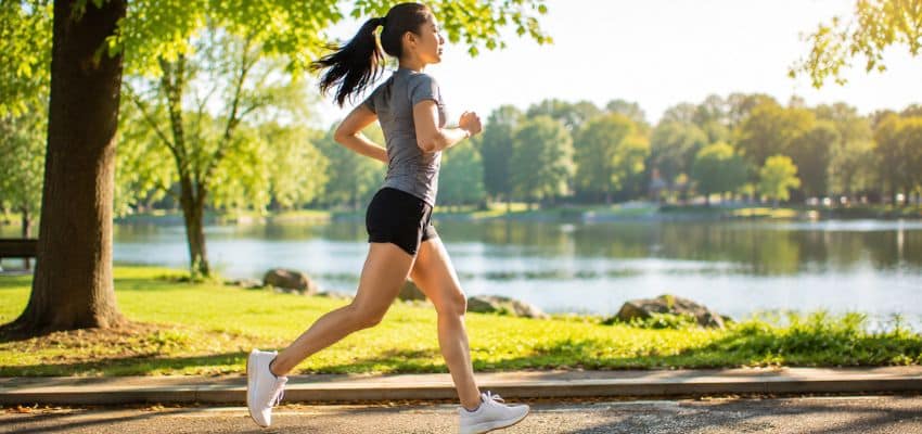 Running Form for Beginners: Build Efficient Habits from Day One featuring a woman practicing efficient running form along a sunny lakeside path in a green park, wearing athletic clothing and white running shoes.